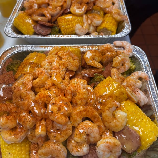 Two foil trays filled with seasoned shrimp, corn on the cob, and potatoes.