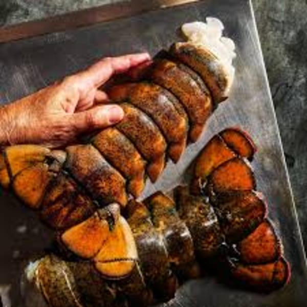 A hand holding a large lobster tail, with several other tails on a metal tray.
