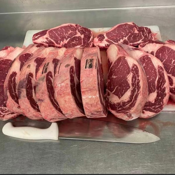 Many raw, marbled steaks are stacked on a white cutting board next to a large butcher knife.