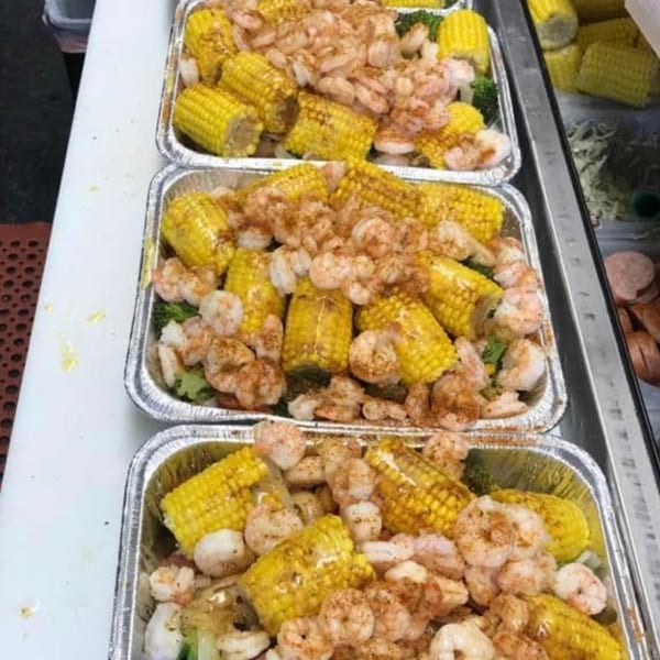 Three foil trays filled with seasoned shrimp, corn on the cob, and broccoli, ready for serving.