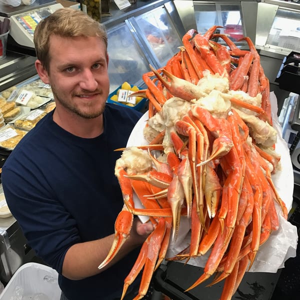 A smiling man in a seafood market holds a large platter piled high with bright orange crab legs.
