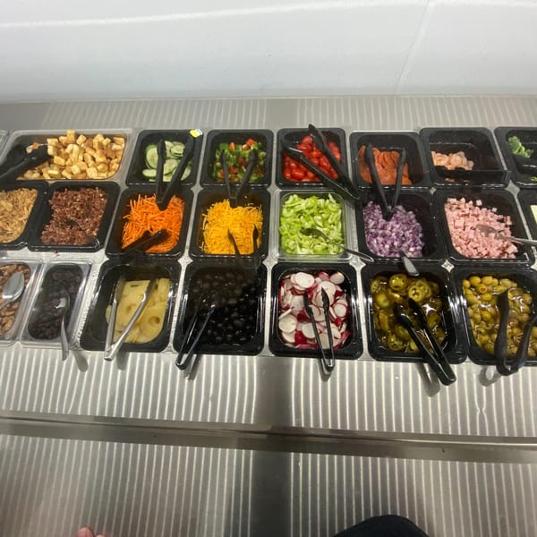A salad bar with various toppings including vegetables, cheeses, and meats in black containers.