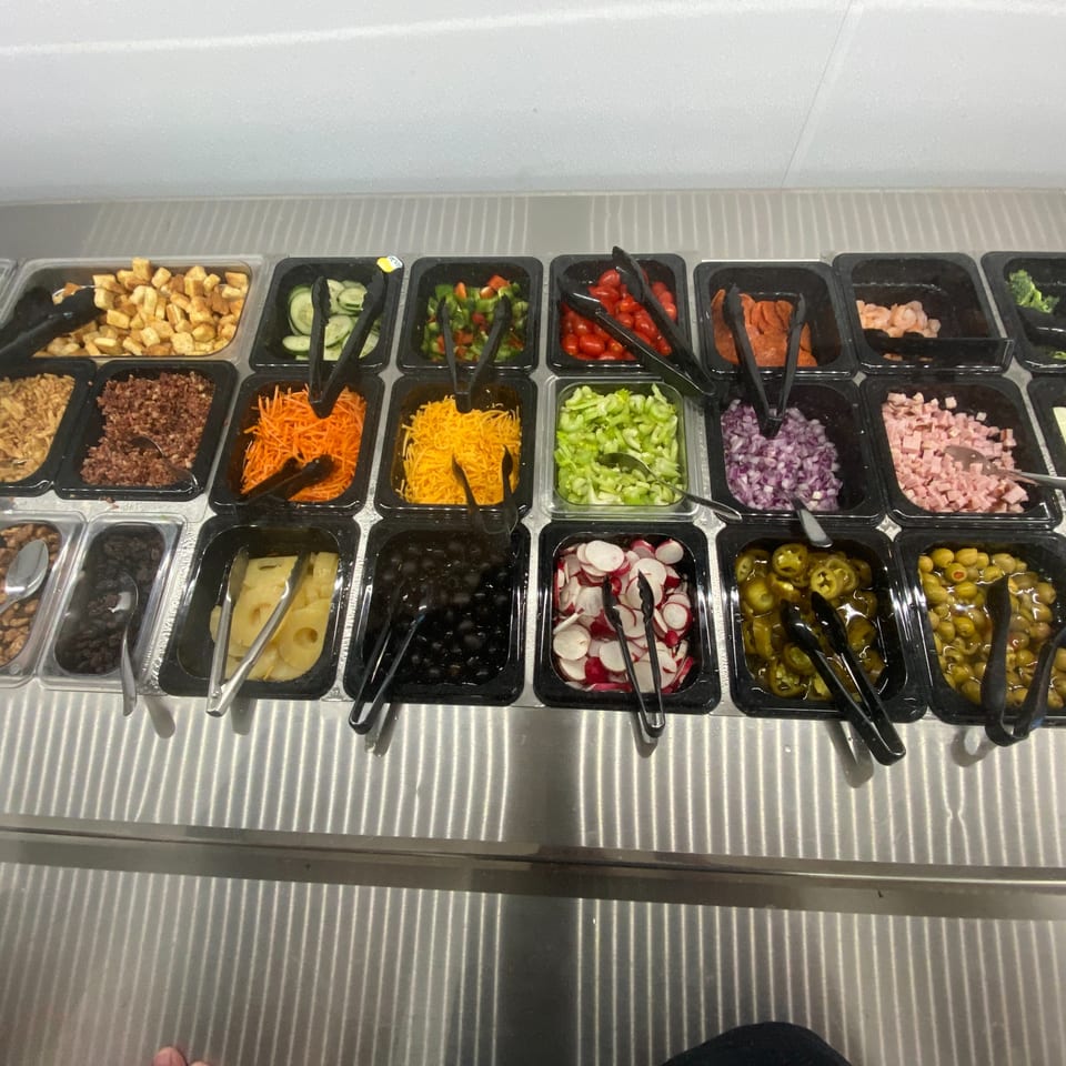 A salad bar with various toppings including vegetables, cheeses, and meats in black containers.